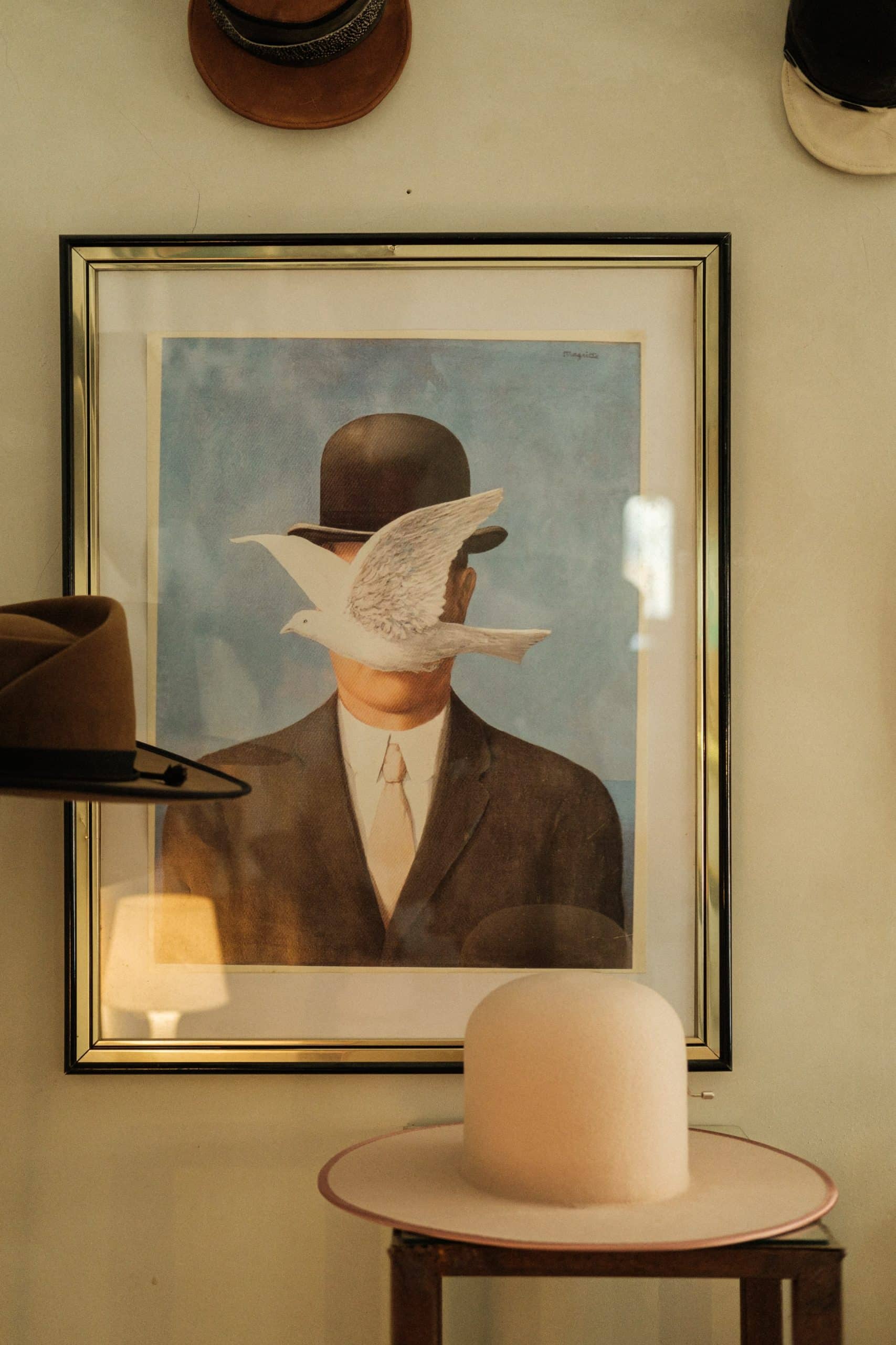 Chapéu claro pousado em cima de mesa de madeira. Quadro na parede com a imagem de um homem de chapéu com uma pomba na frente do rosto.
