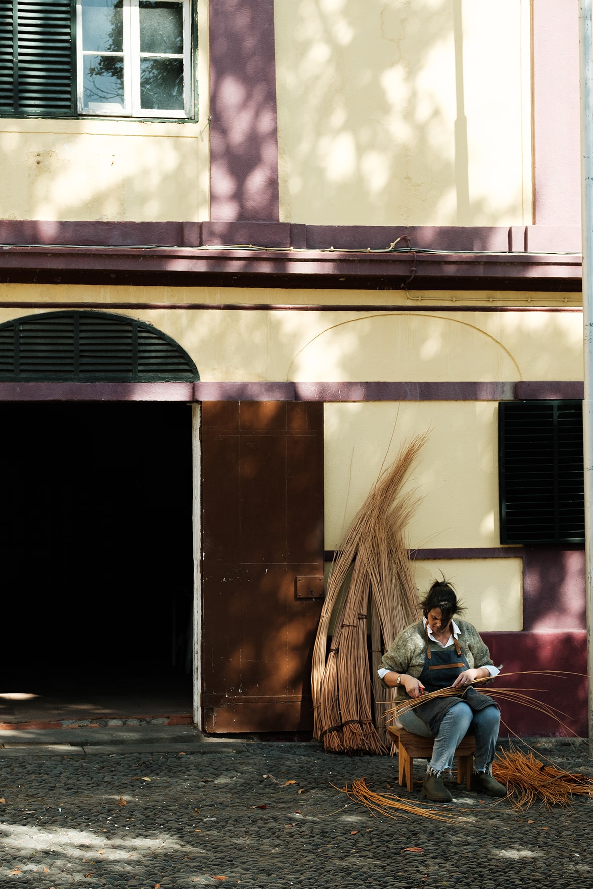 Catarina de Jesus a trabalhar fora do atelier, sentada numa cadeira com molhos de vime atrás, encostados à parede.