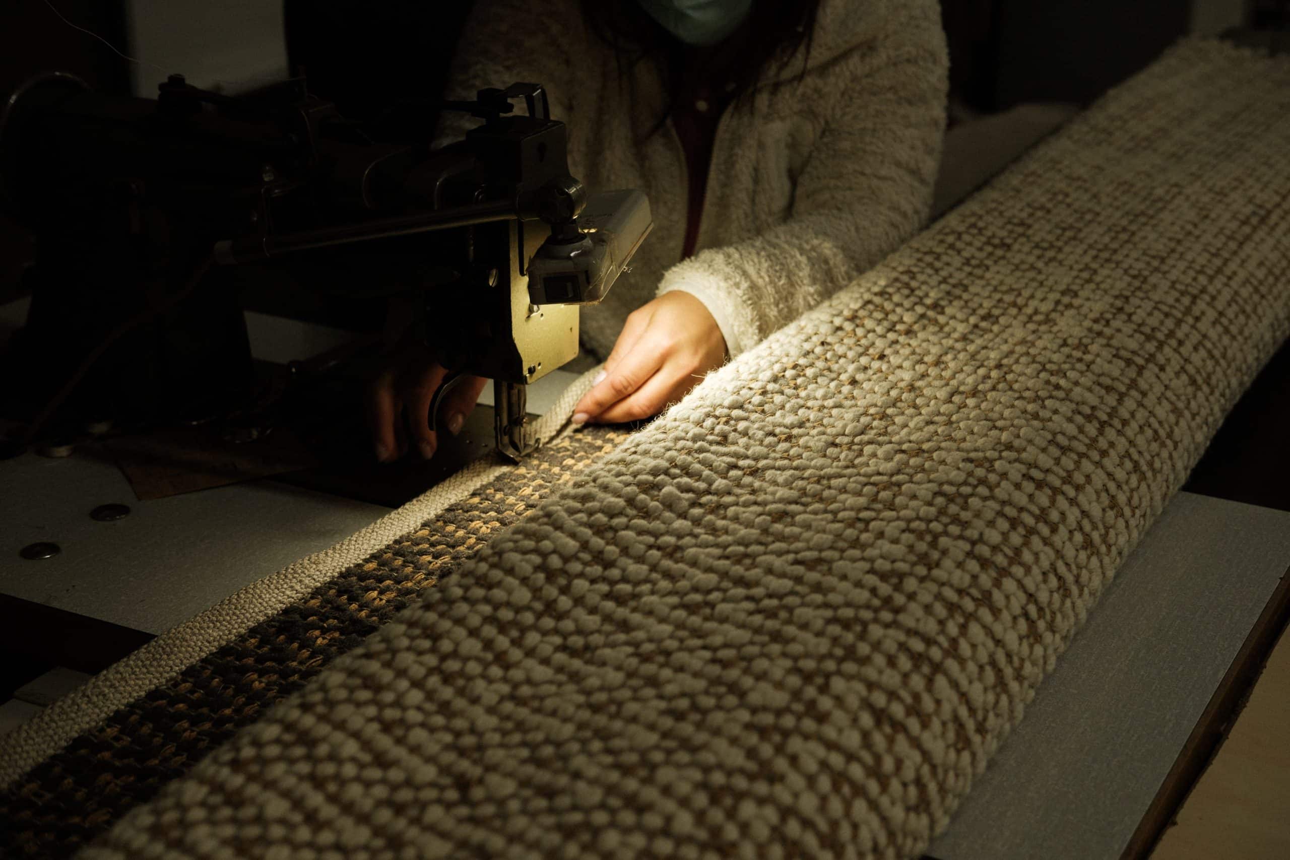 Mãos costurando um tapete na máquina de costura, com uma luz suave acima.