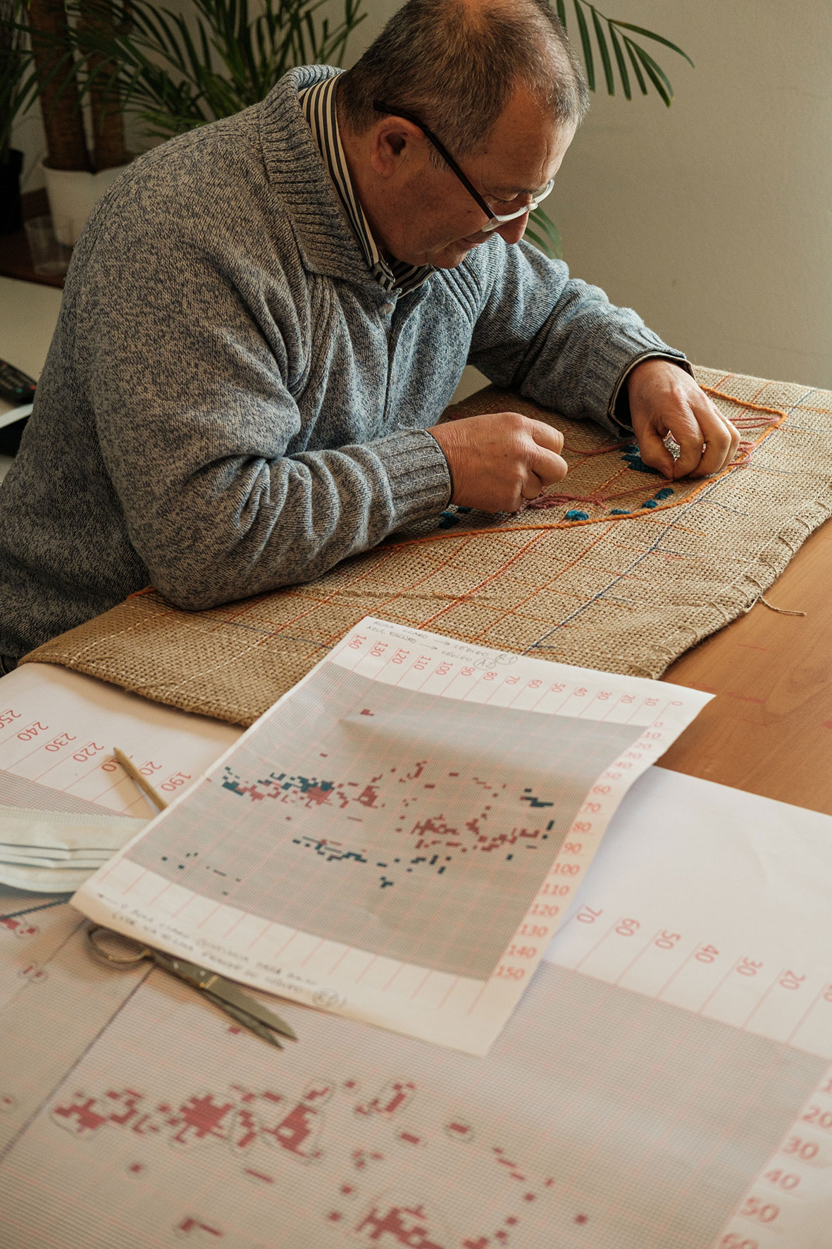 Pai da artista sentado, borda fio cor de laranja numa tela de serapilheira. Ao seu lado, folhas de planeamento e esboço.