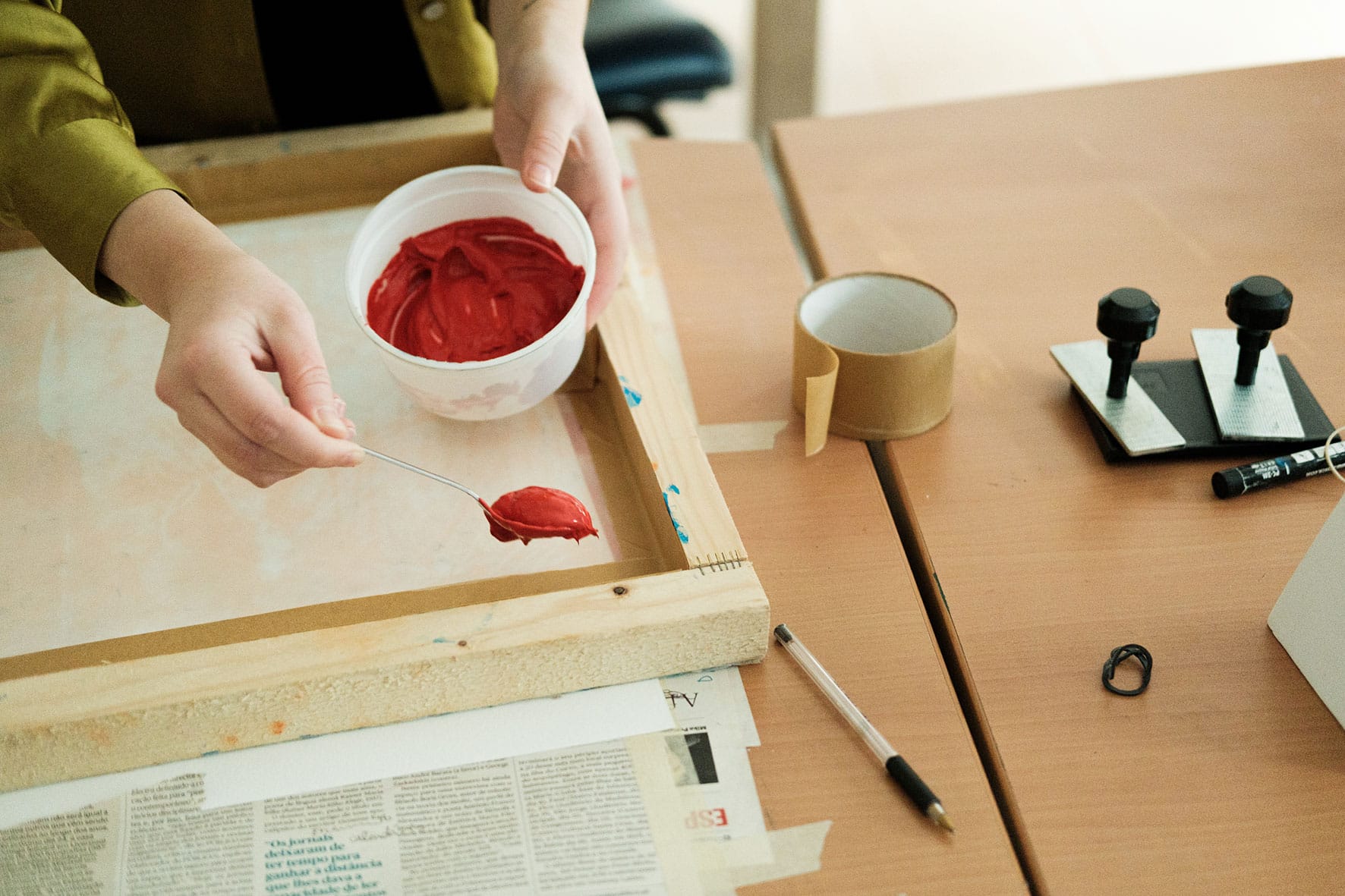 Artista coloca tinta vermelha na superfície de tela, com o auxilio de colher.
