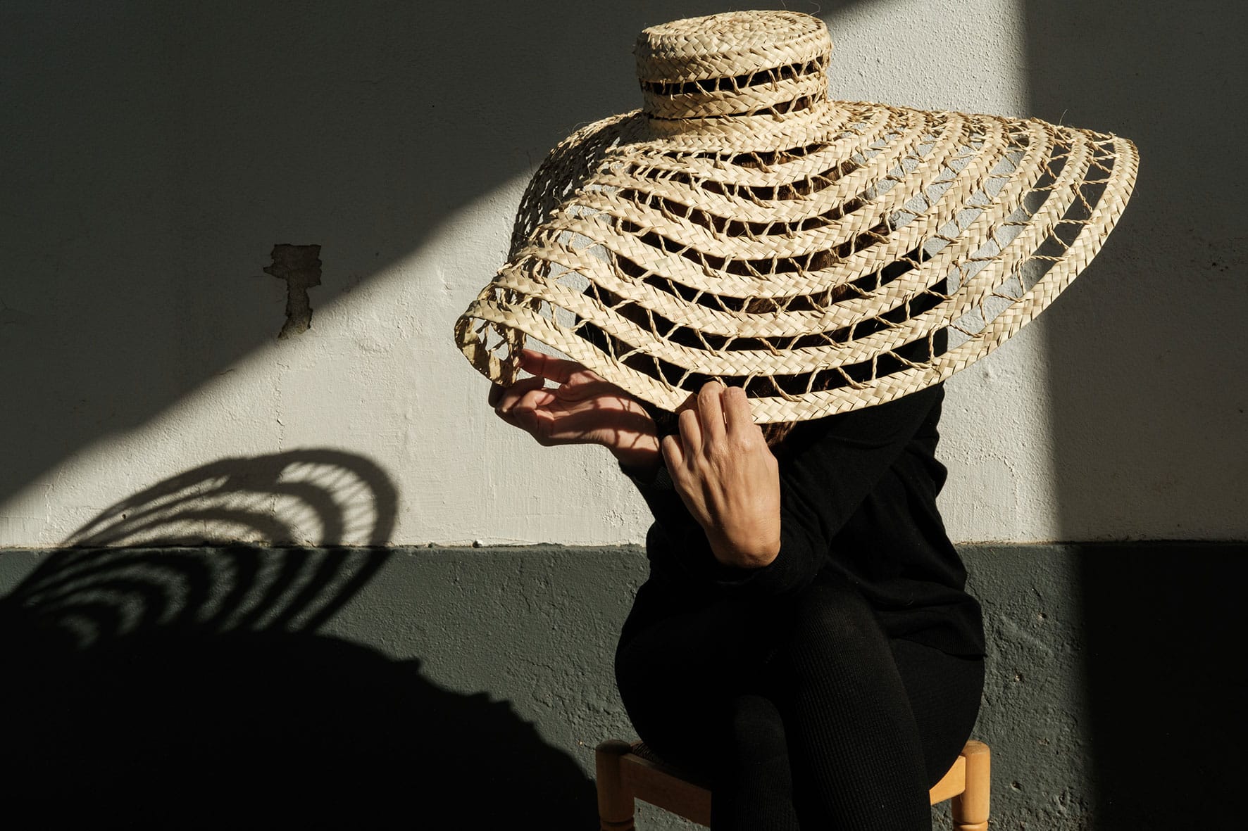 Artista usa chapéu de grande dimensão feito de palma. A sua sombra incide na parede.