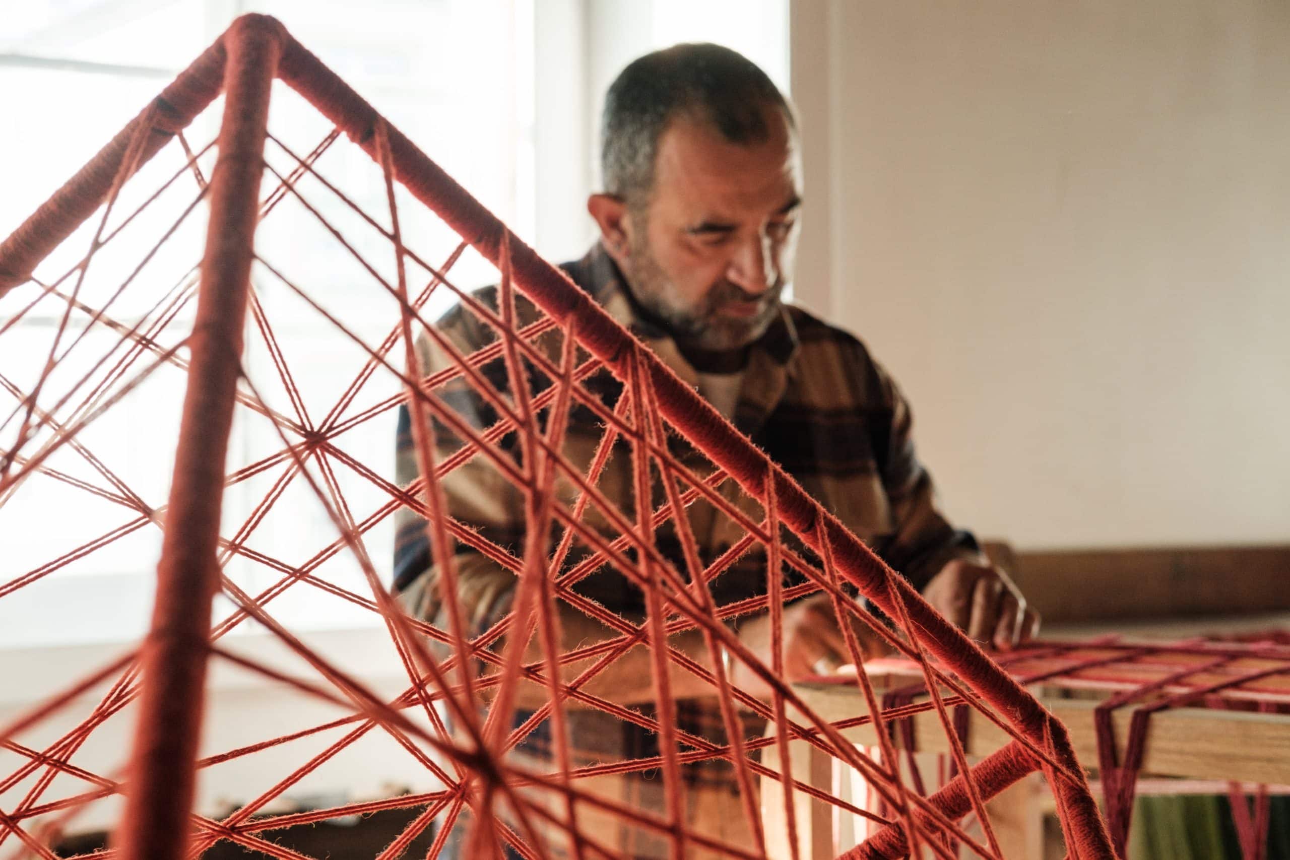 Estrutura cúbica tecida a fios de lã vermelhos em primeiro plano e João Bruno no fundo, trabalhando no assento de cadeira.