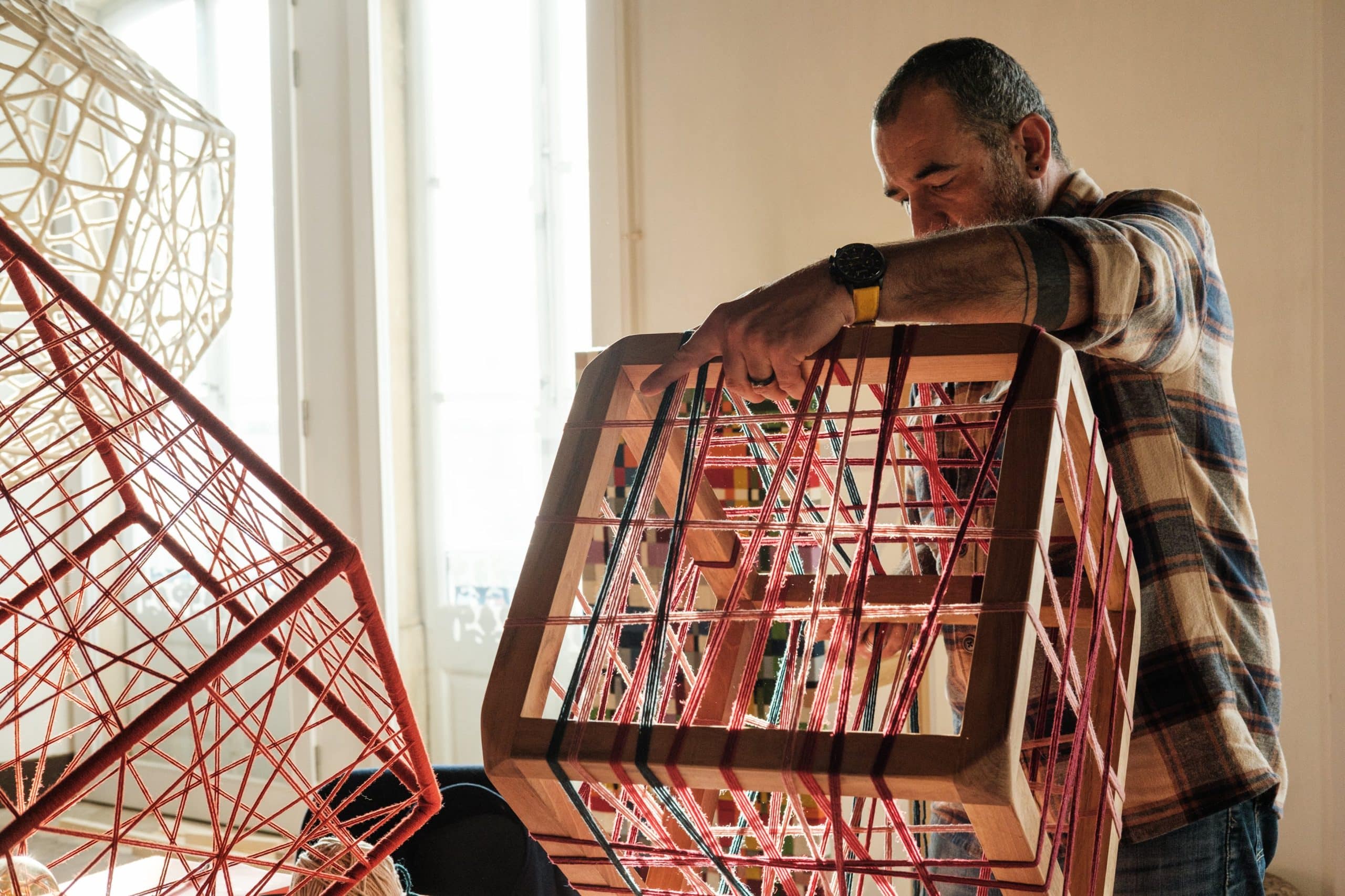 Artesão dá acabamentos em estrutura cúbica de madeira, tecendo fios vermelhos.