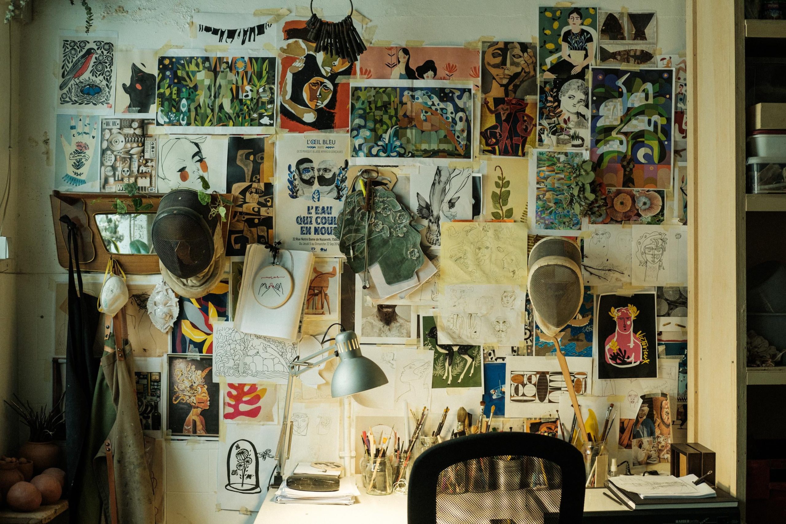 Espaço de trabalho, mesa e cadeira voltadas para uma parede repleta de posters, papéis, fotografias, ilustrações, etc.