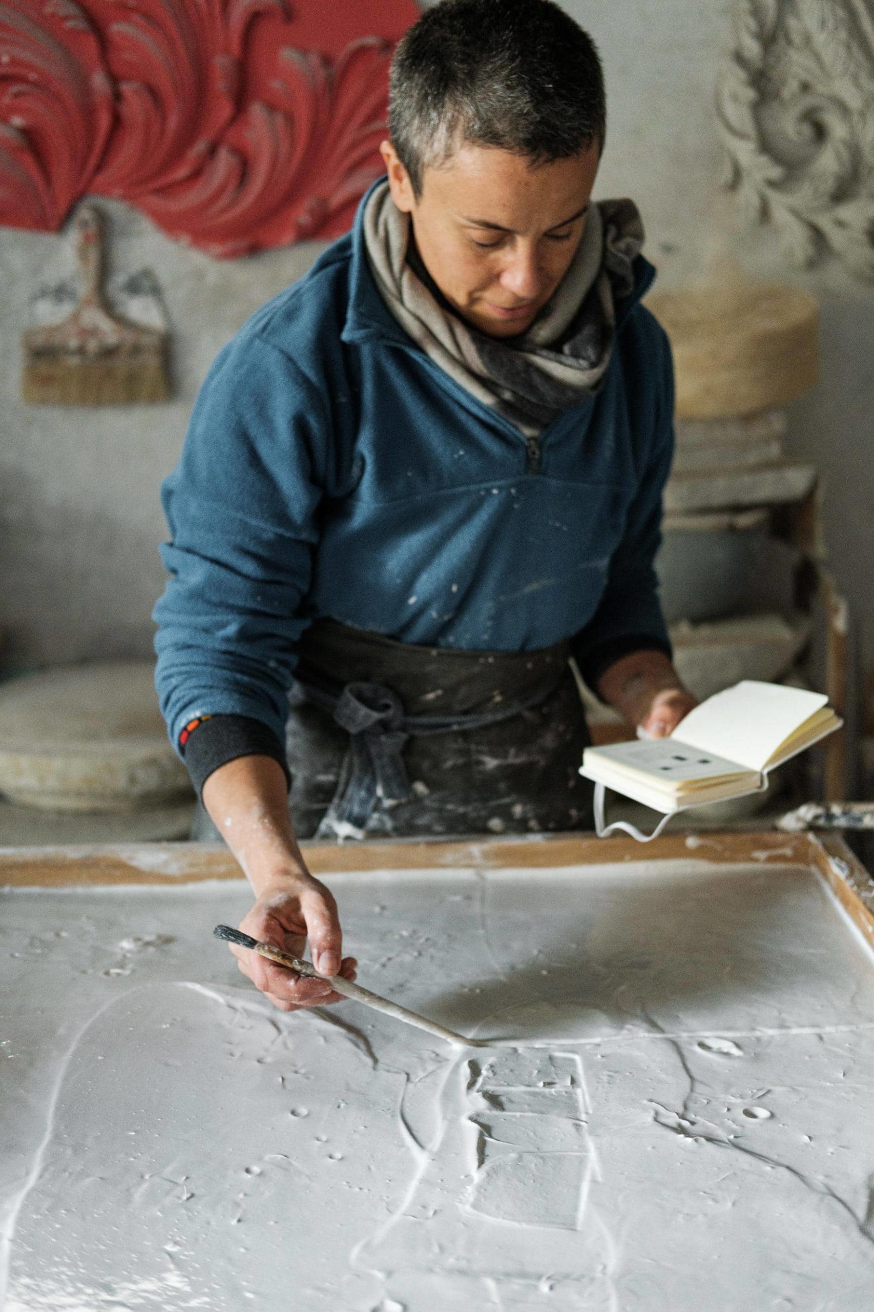 Artista Iva Viana molda placa de gesso com uma mão de acordo com o esboço que tem na outra.
