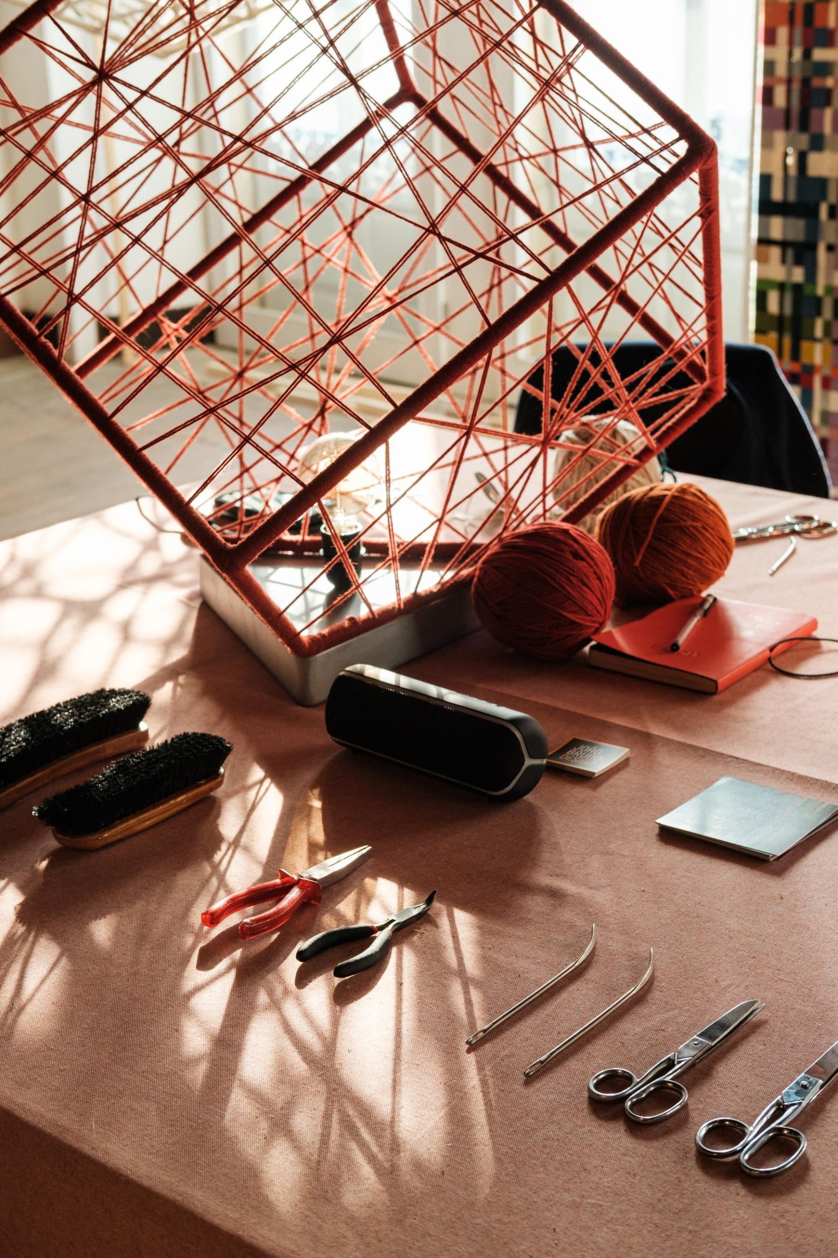 Mesa de trabalho com diferentes materiais, com foco na peça de madeira cúbica envolvida em fios de lã vermelhos.