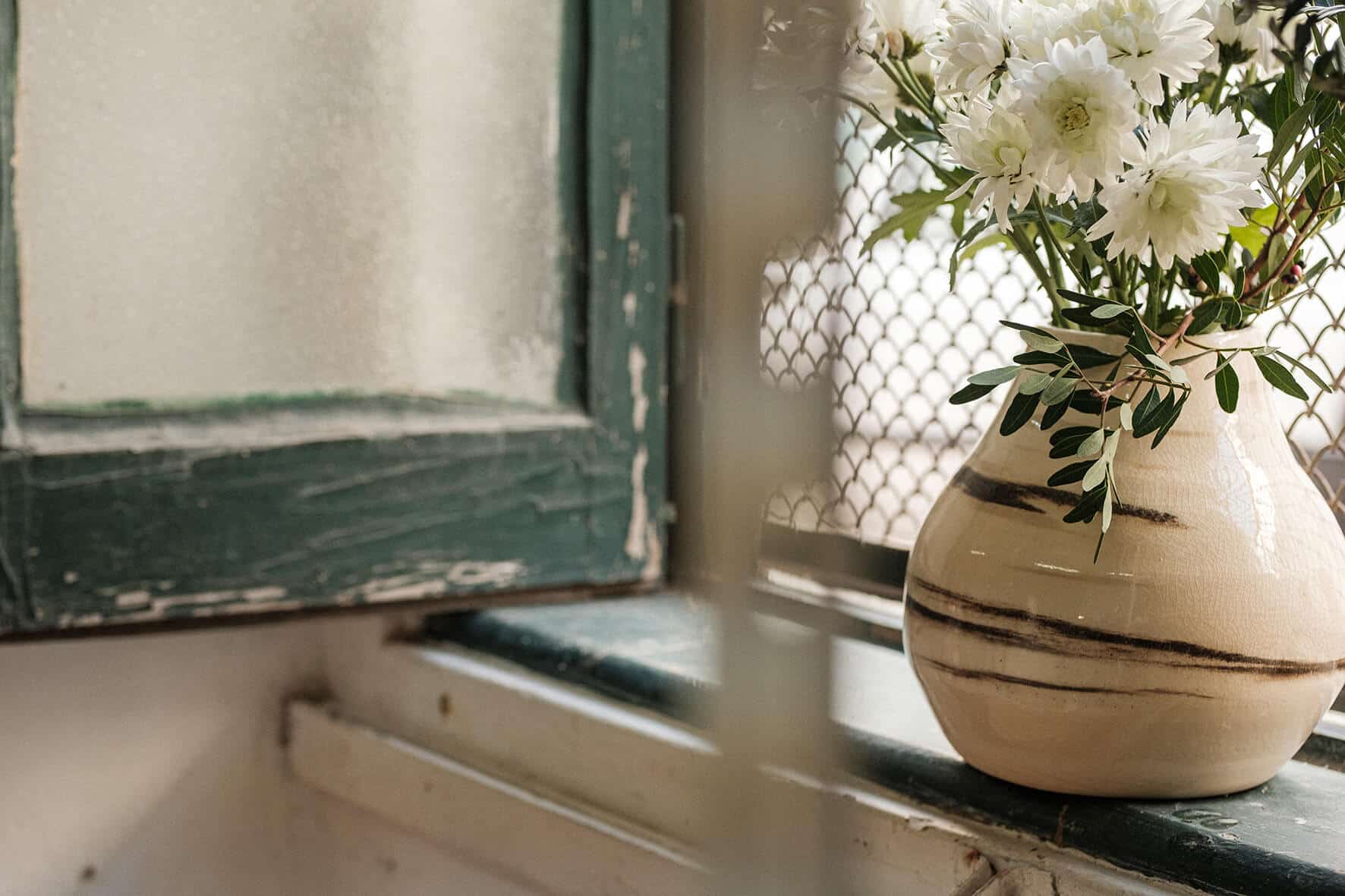 Vaso de cerâmica branco com flores, pousado no parapeito da janela.