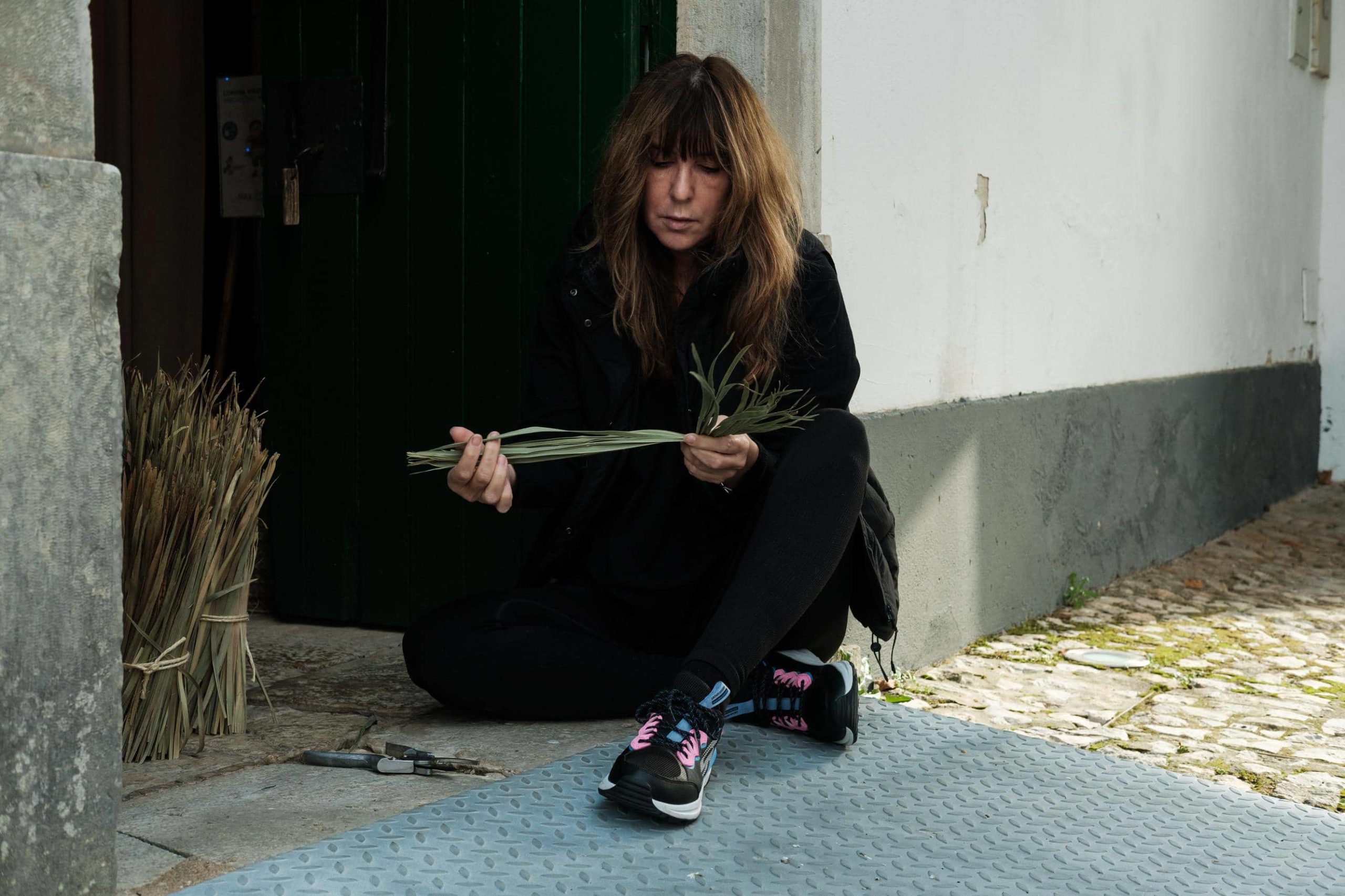 Artista Maria João sentada no chão à porta a segurar em palma destrançada.