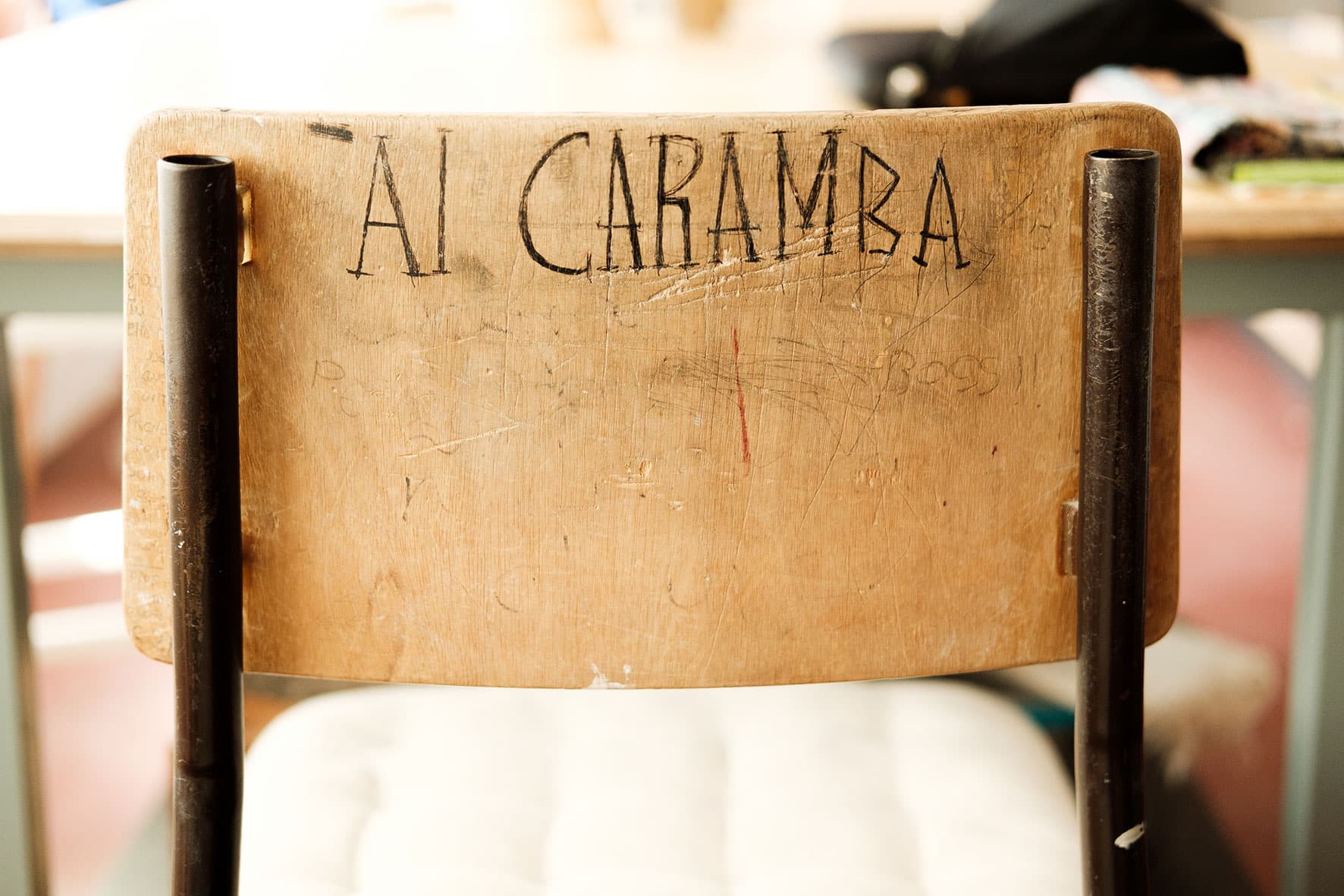 Encosto de cadeira de madeira, onde se encontra escrito manualmente a frase "Ai Caramba" em letras maiúsculas.
