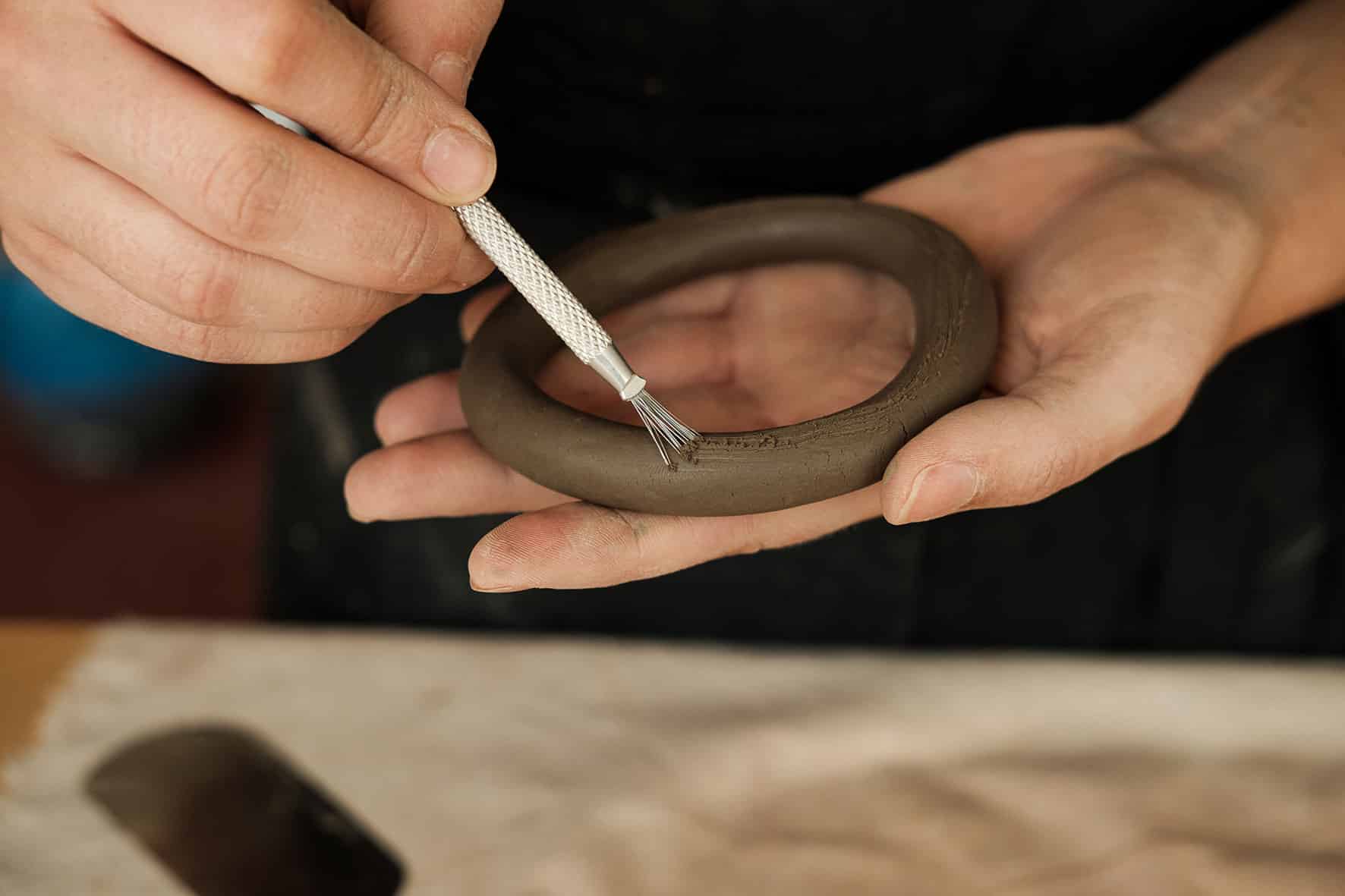 Mão de ceramista segura um rolo moldado em forma circular enquanto passa levemente uma pasta com ajuda de pincel.